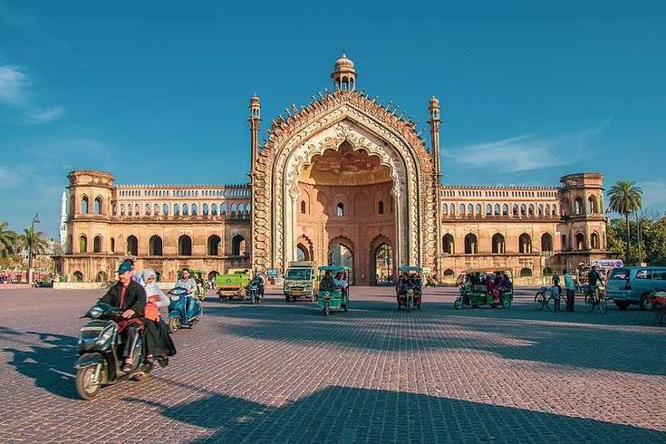 A wide-angle view of the Rumi Darwaza in Lucknow, India, under a clear blue sky. The ornate, grand archway dominates the centre of the image. The street in front is a busy scene with various vehicles. The tourist place in Lucknow includes motorbikes, rickshaws, and cars, moving about, along with a few pedestrians. The sunlight casts shadows on the ground and highlights the detailed architecture of the gateway.