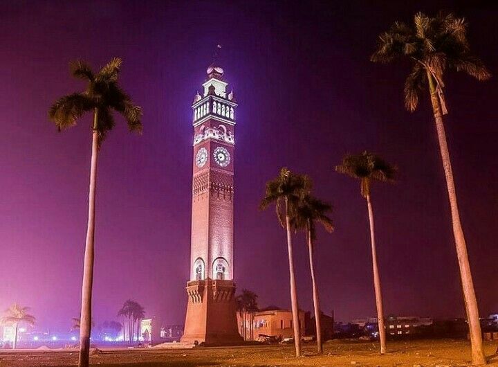 A tall, ornate brick clock tower stands illuminated at night against a dark purple sky. The tower has multiple clock faces and is topped with a decorative spire. The tourist place in Lucknow Several tall palm trees are scattered in the foreground and around the base of the tower.